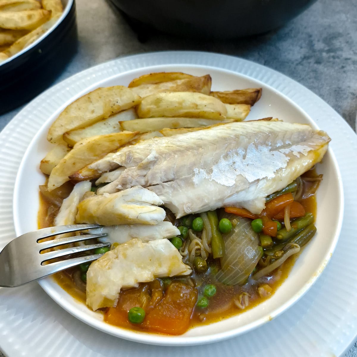 A sea bass fillet on a plate with garlic sauce and french fries, with the skin removed. The fish is being flaked with a fork.