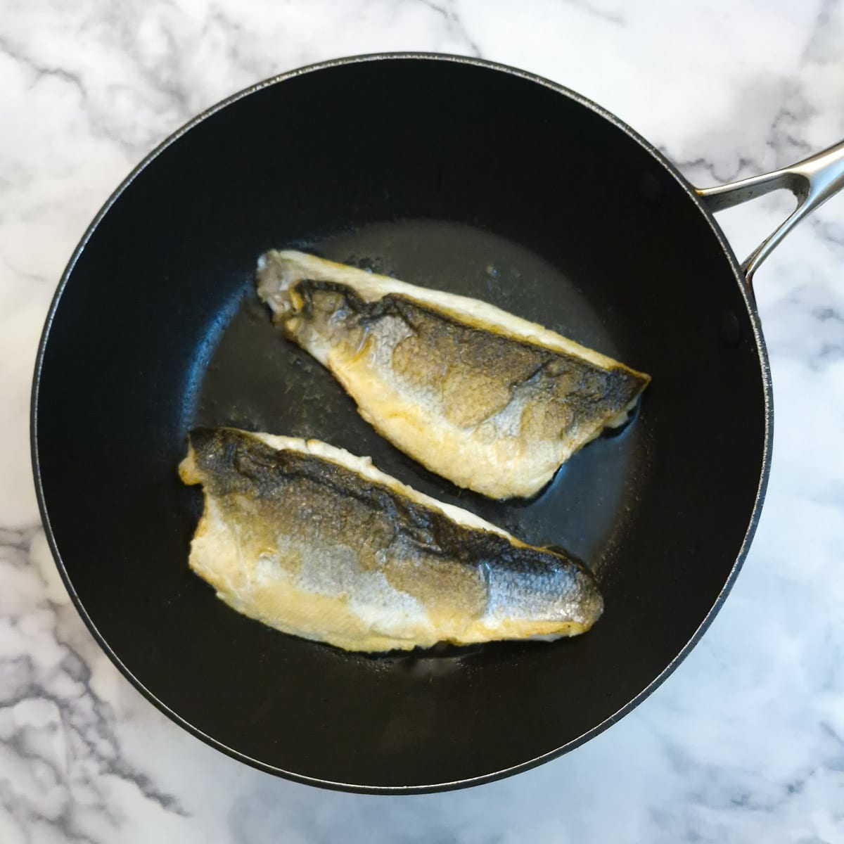 Two pieces of sea bass browning in a frying pan.