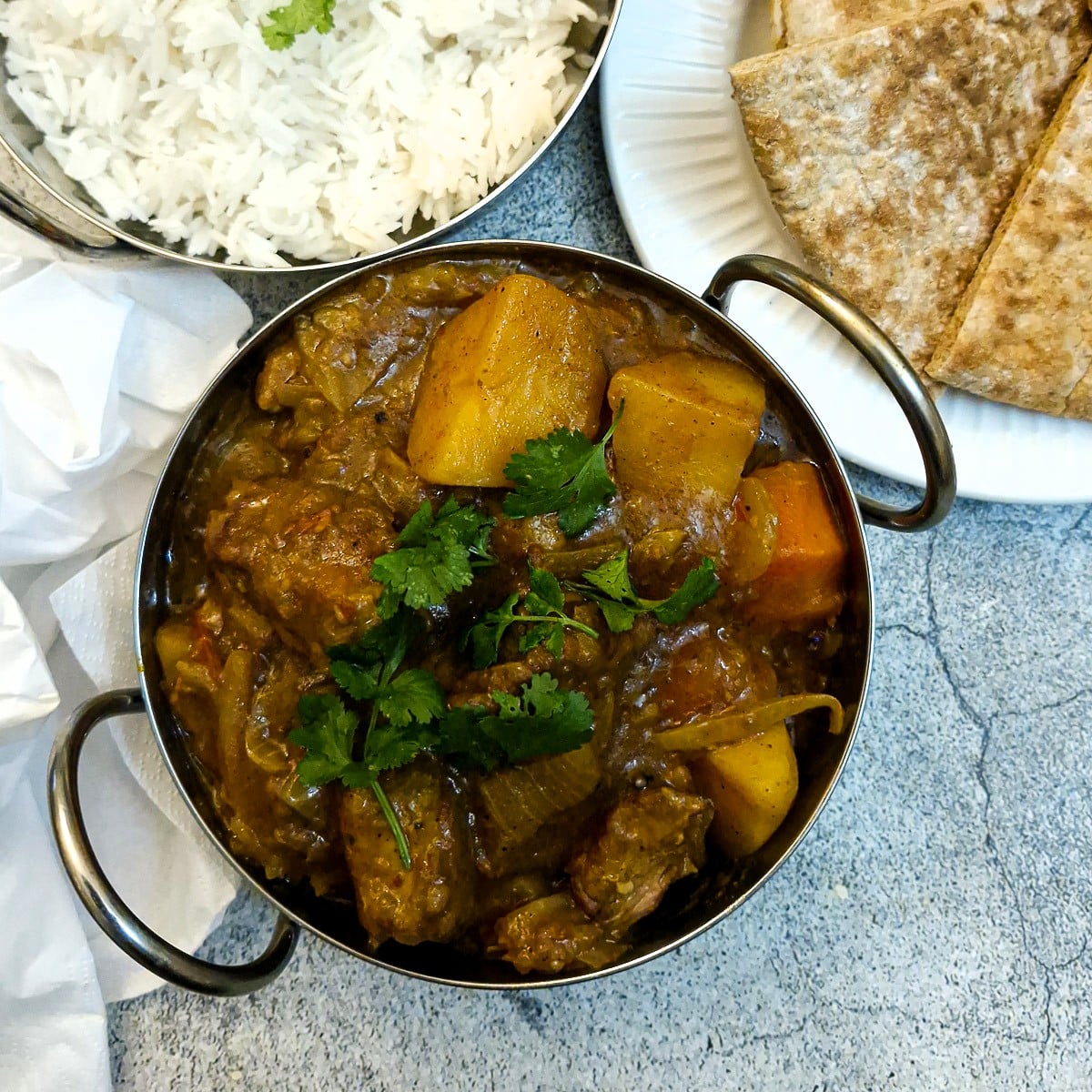 A balti dish of slow cooker lamb curry. There is a dish of rice and a plate of naan bread alongside.