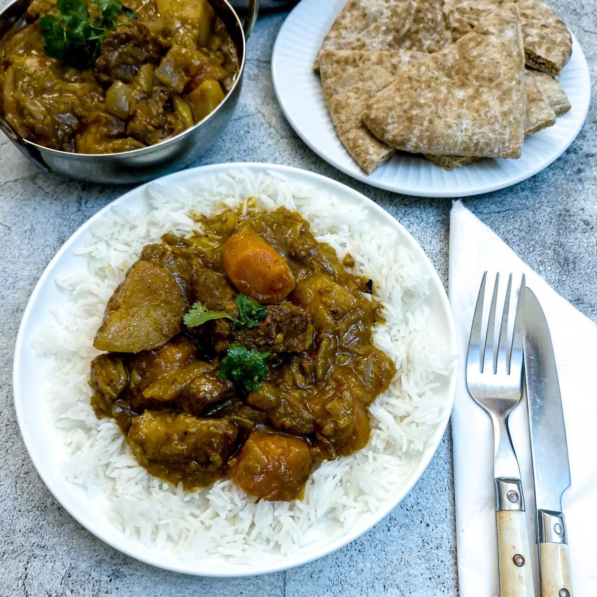 Lamb ccurry on a bed of white rice on a white plate on a table with a knife and fork. There is a plate of naan bread alongside.