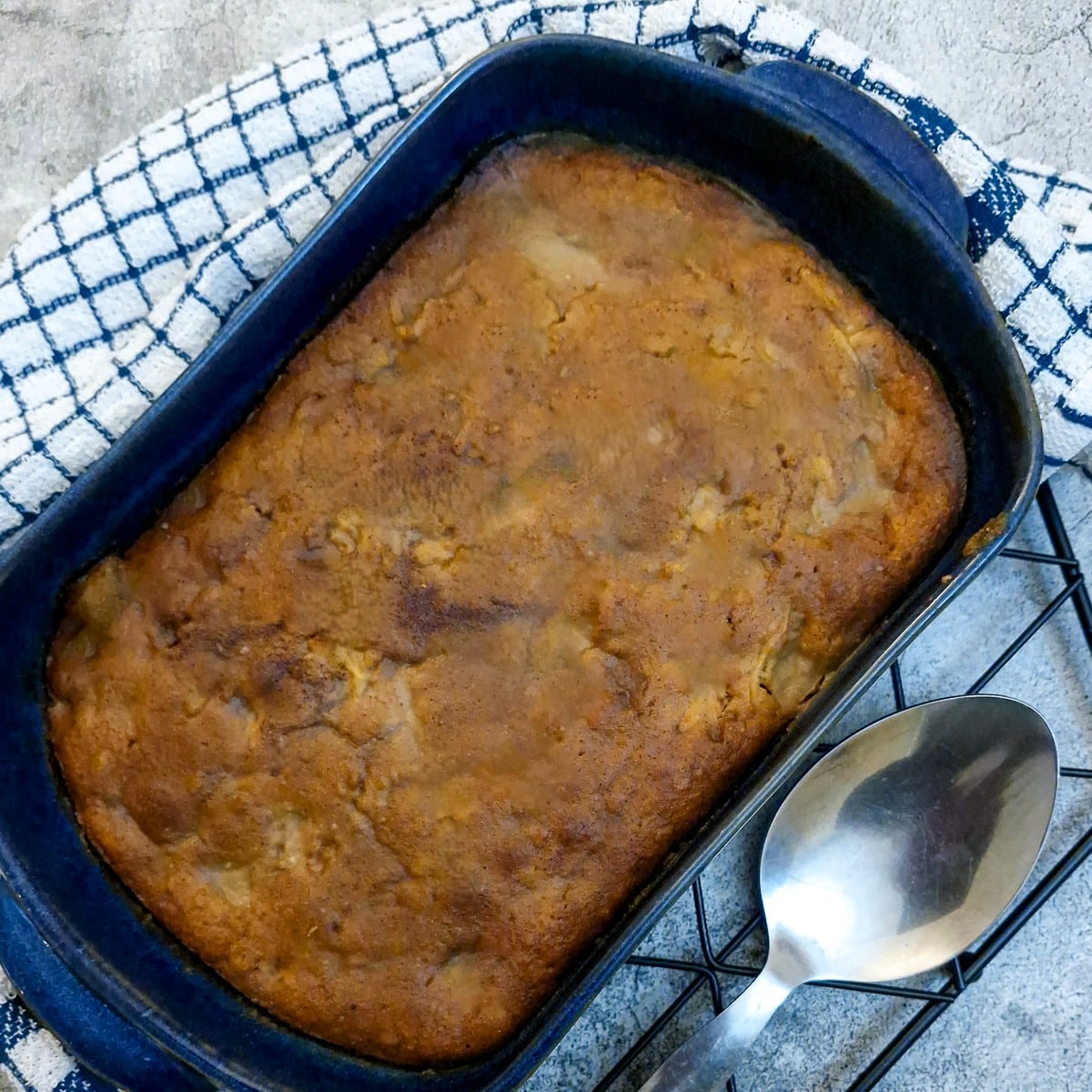 Overhead shot of a blue baking dish of Biscoff apple pudding cake. There is a large serving spoon next to the dish along with a blue-checked tea towel.