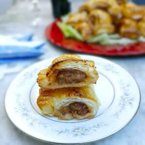 Two pork and cranberry sausage rolls on a small white plate. There is a dish of sausage rolls in the background.