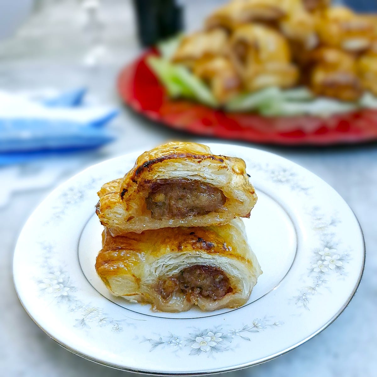 Two pork and cranberry sausage rolls on a small white plate. There is a dish of sausage rolls in the background.