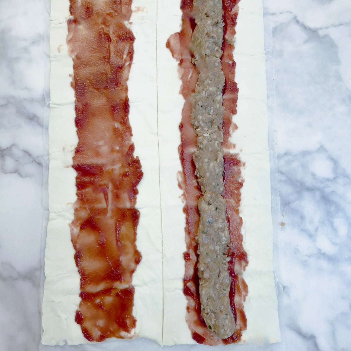 2 lenghts of puff pastry, both topped with a layer of cranberry sauce. There is a roll of pork sausage meat on top of the cranberry sauce on one of them.