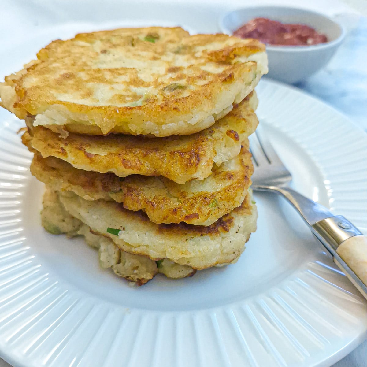 A pile of potato cakes on a white plate. There is a small dish of tomato ketchup in the background.