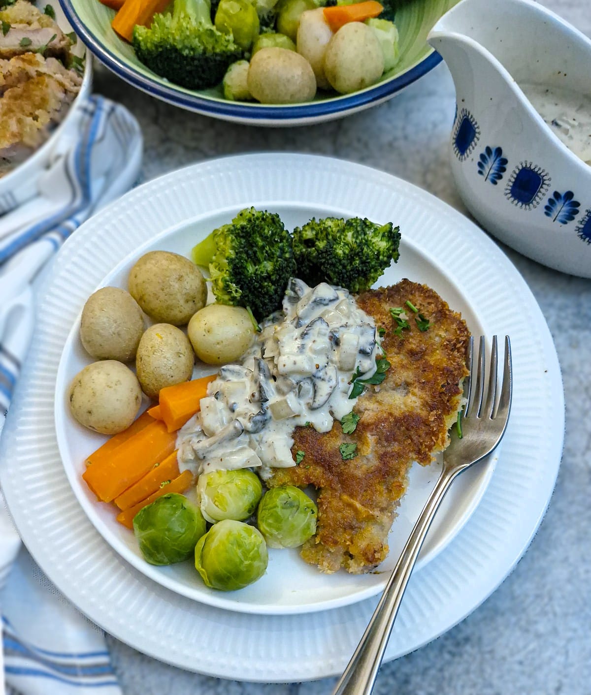 A crumbed pork chop on a plate with vegetables and potatoes. The chop has been partially covered with creamy mushroom mustard sauce.