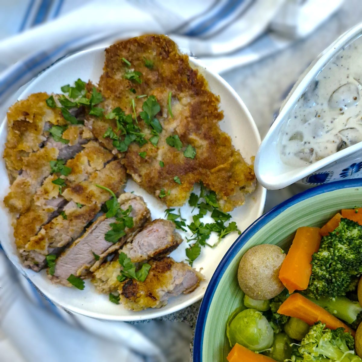 Crumbed pork chops on a plate, garnished with parsley. There is a jug of mushroom mustard sauce alongside.