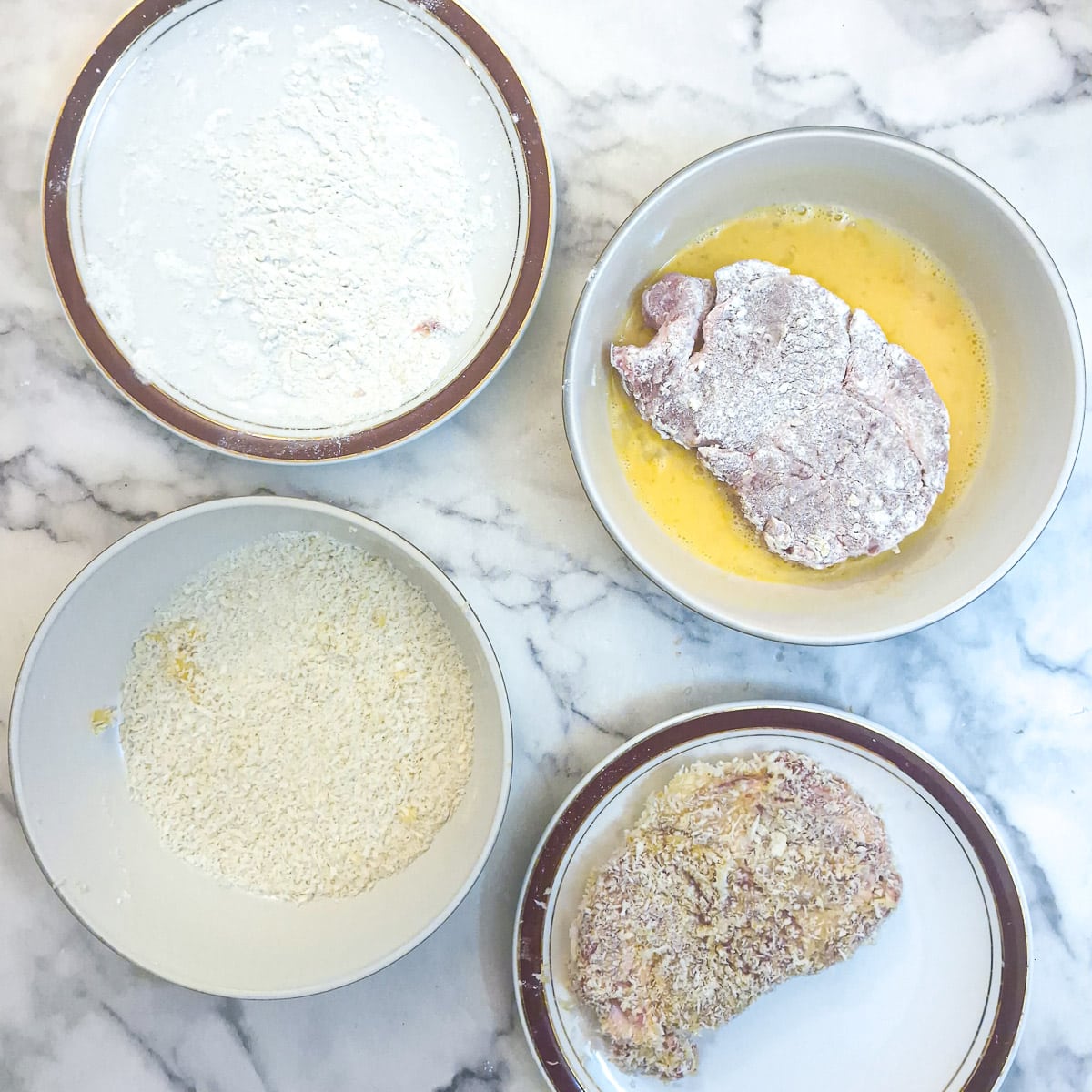 4 plates and dishes, 3 of them holding flour, beaten egg and panko breadcrumbs. The 4th plate holds a crumbed pork chop.