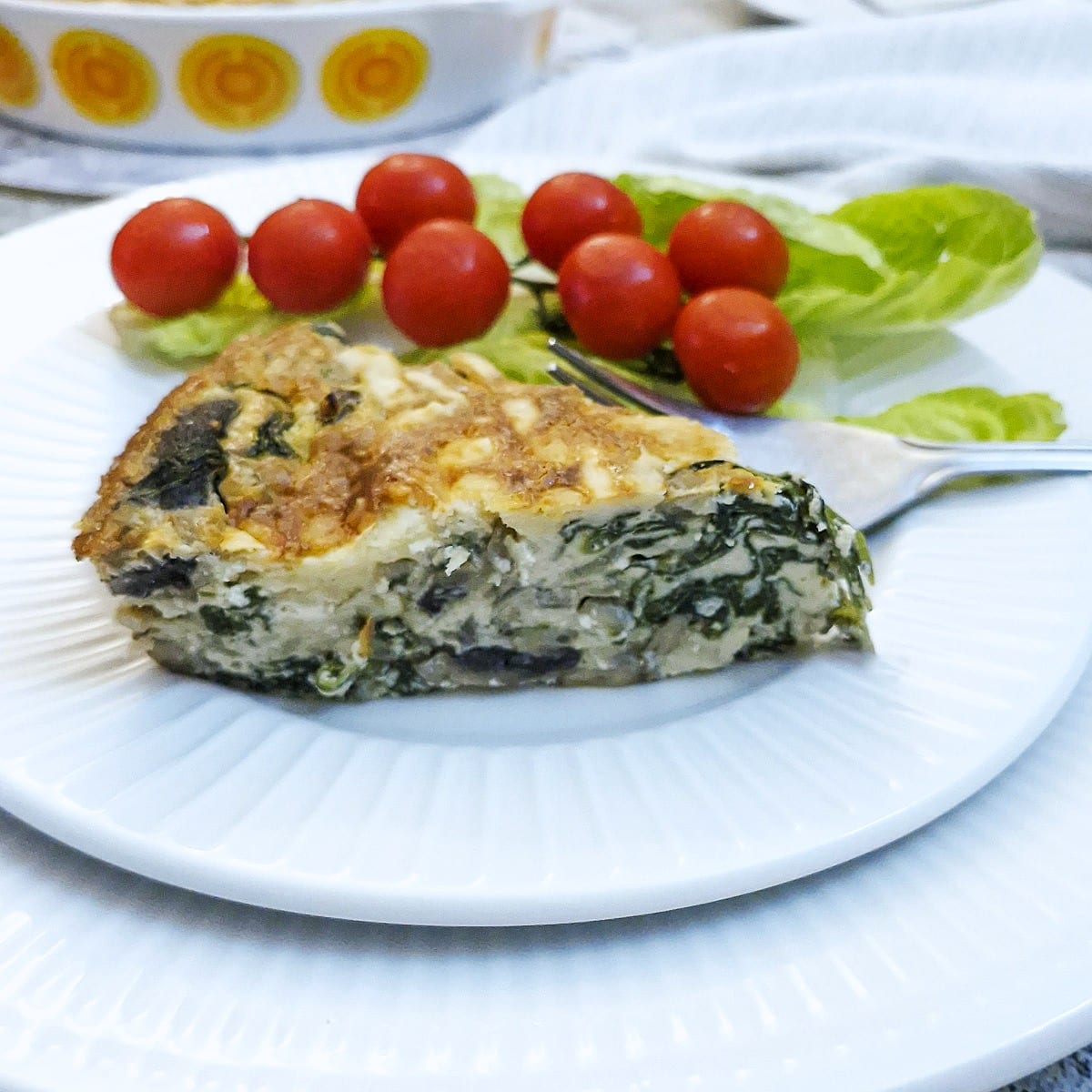 A slice of crustless spinach quiche on a white plate, showing the firm texture of the quiche. There are a few cherry tomatoes and slices of lettuce on the side.