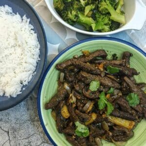 A dish of cumin beef stir-fry with dishes of rice and broccoli alongside.