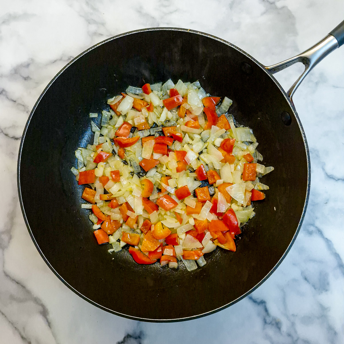 Chopped red peppers and onions softening in a black frying pan.