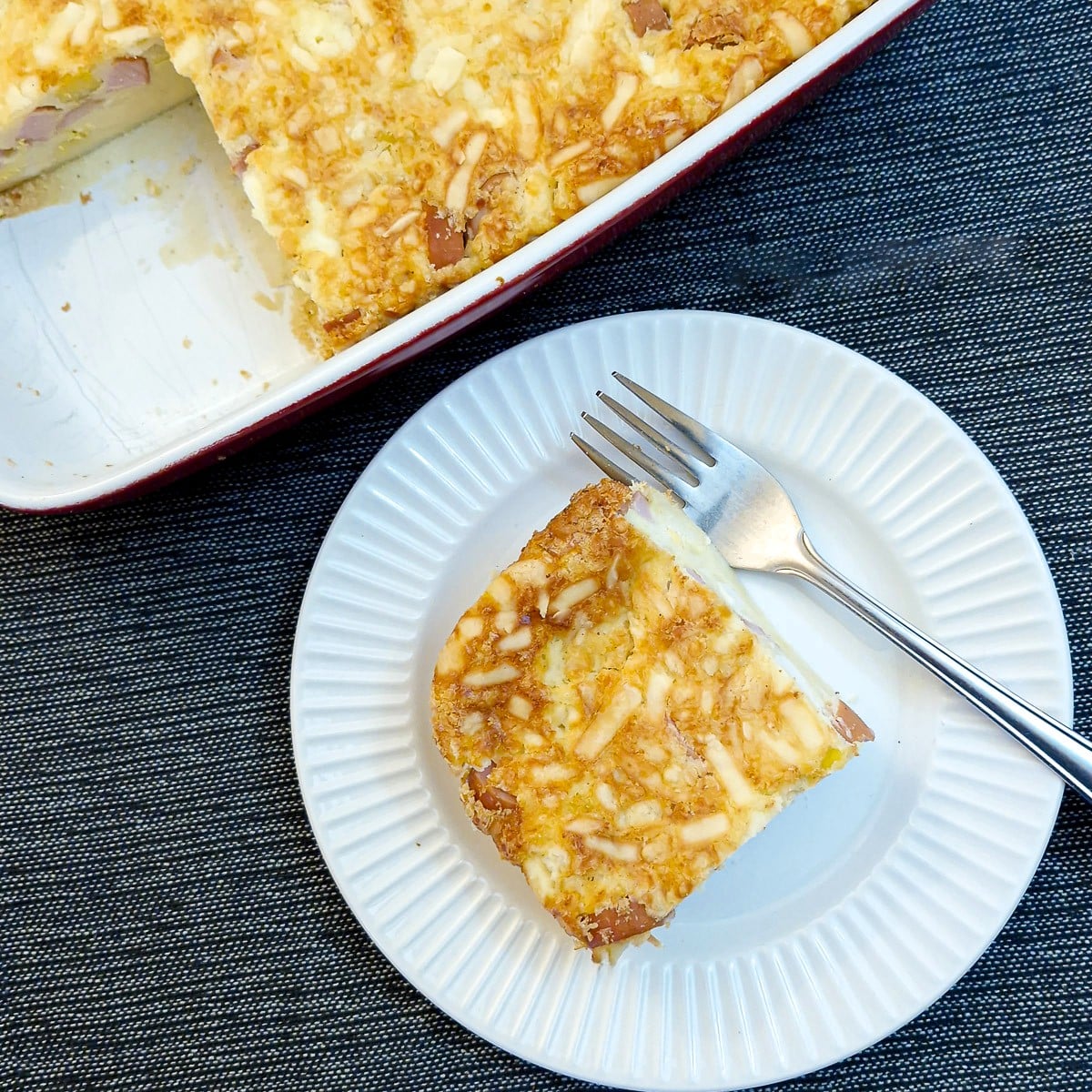 A slice of crustless savoury tart on a small white plate with a cake fork. There is a baking dish of the tart in the background.