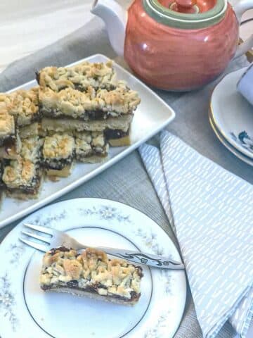 A tea tray laid with a plate of date and walnut crumble bars. A teapot, 2 cups and saucers and a slice of the date bar on a plate are also on the tray.