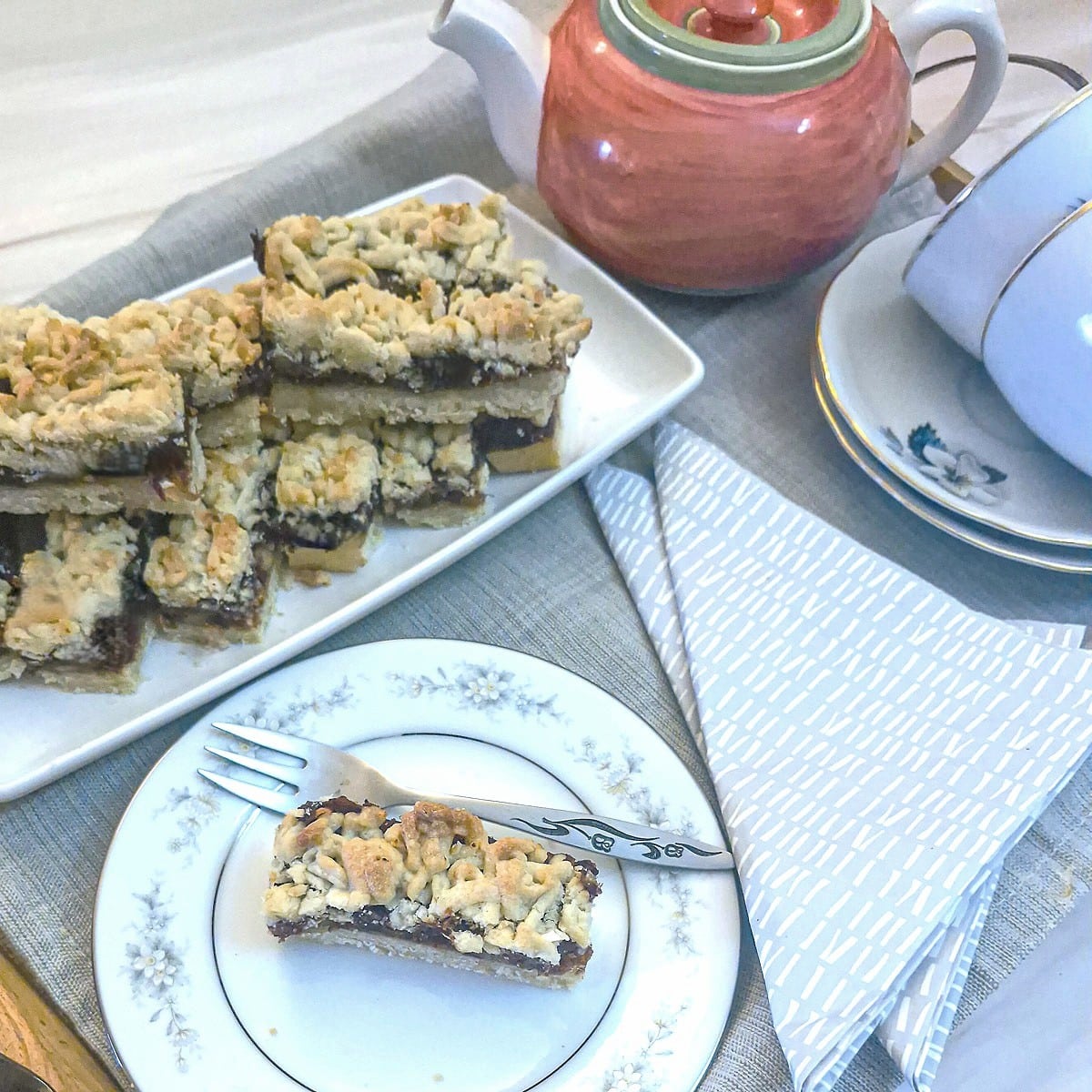 A tea tray laid with a plate of date and walnut crumble bars.  A pink teapot, 2 cups and saucers and a slice of the date bar on a plate are also on the tray.