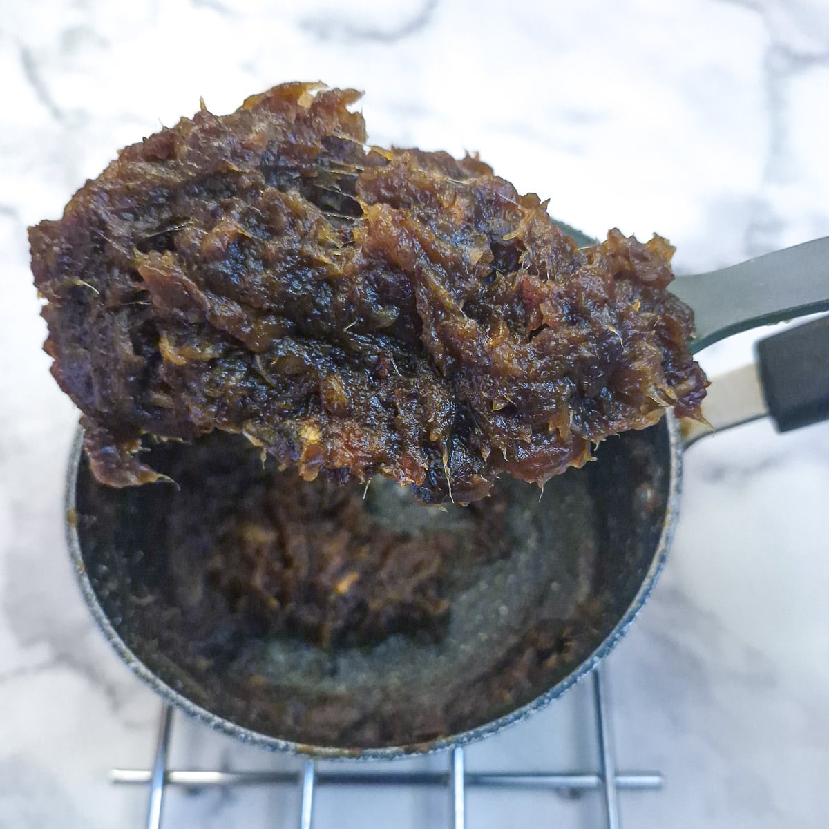 A large spoon ful of date paste after it has boiled. The spoon is held above the pan.