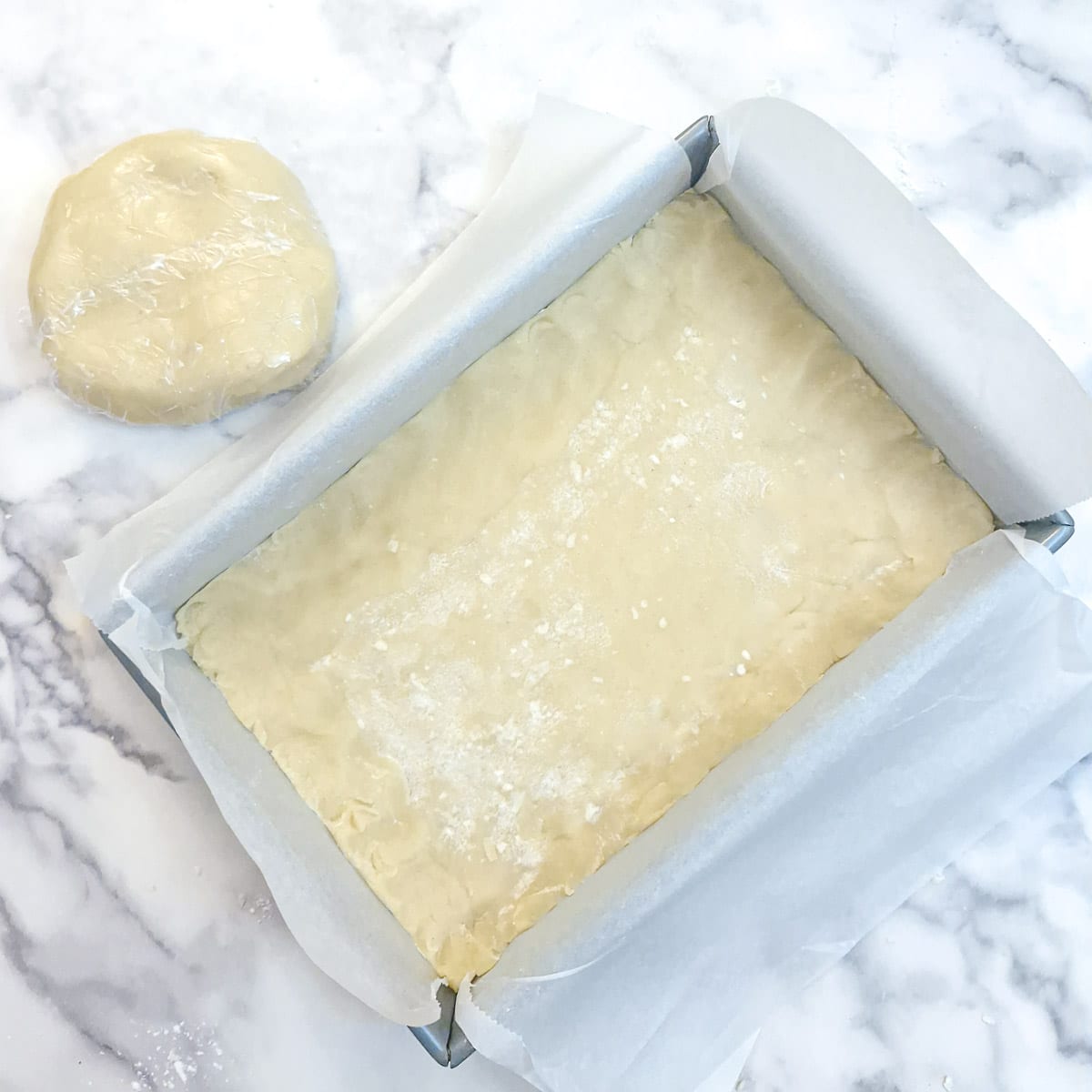 A baking tray lined with baking parchment with the dough pressed onto the base. There is a lump of dough at the side of the baking tray which will be wrapped in plastic and chilled in the freezer.