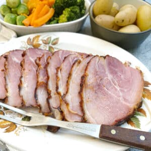 Slices of ham on a serving plate with a serving fork. There are dishes of potatoes and vegetables in the background.