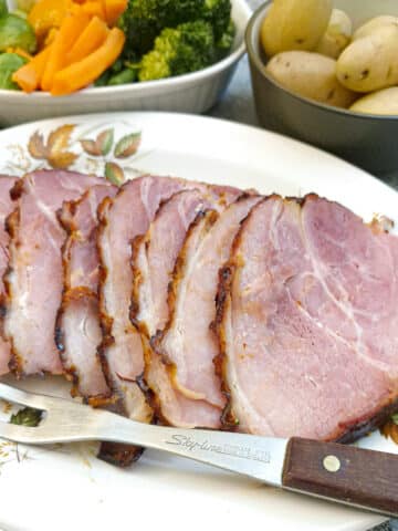 Slices of ham on a serving plate with a serving fork. There are dishes of potatoes and vegetables in the background.