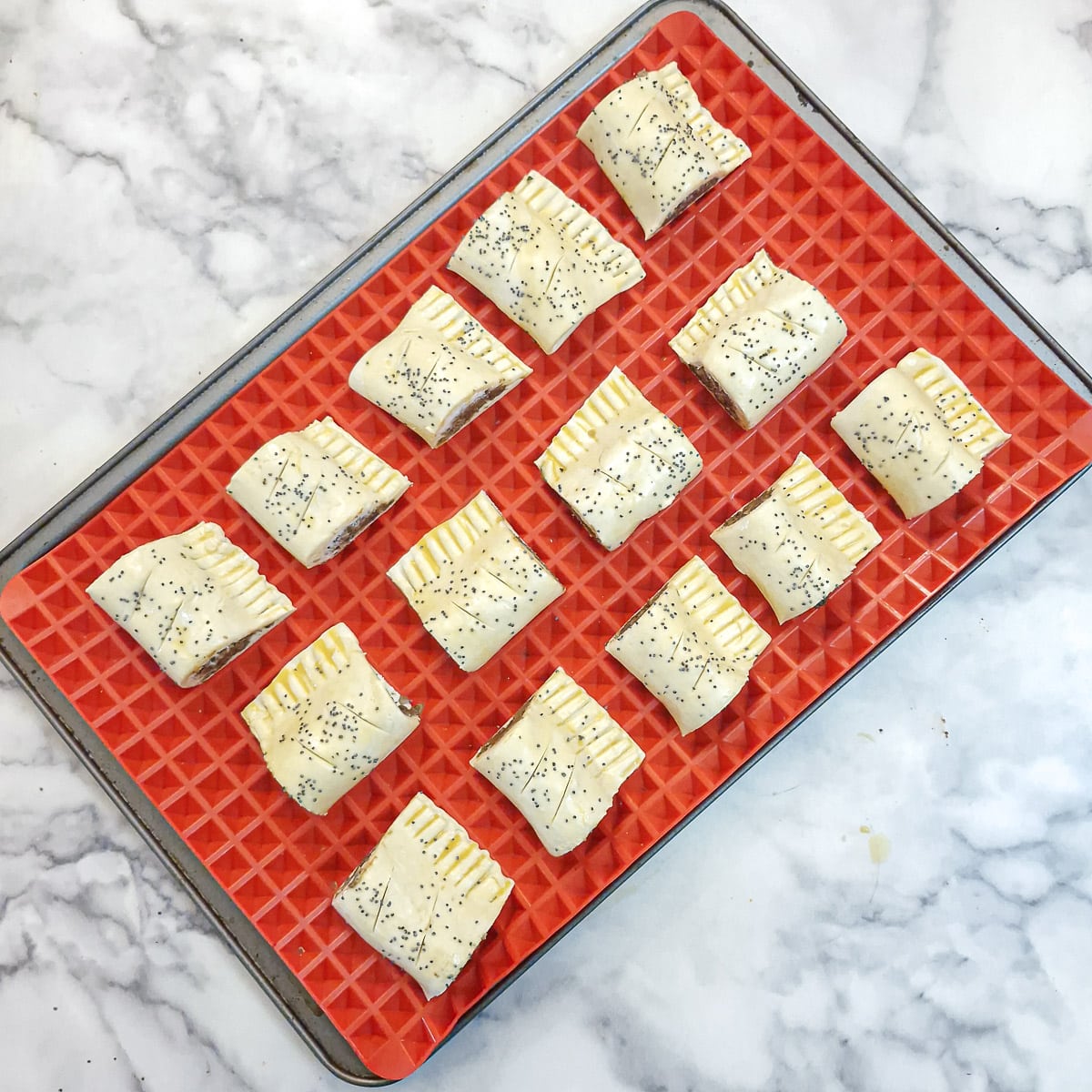 14 unbaked sausage rolls on a red silicone-lined baking tray.