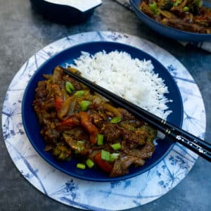 A blue dinner plate with a helping of rice and sweet chilli beef stirfry. There is a pairof black and gold chopsticks balanced on the plate..