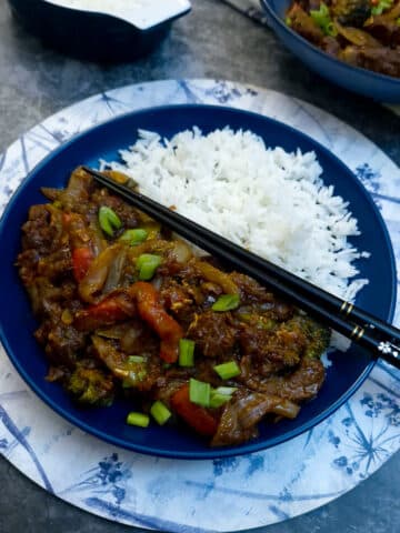 A blue dinner plate with a helping of rice and sweet chilli beef stirfry. There is a pairof black and gold chopsticks balanced on the plate..