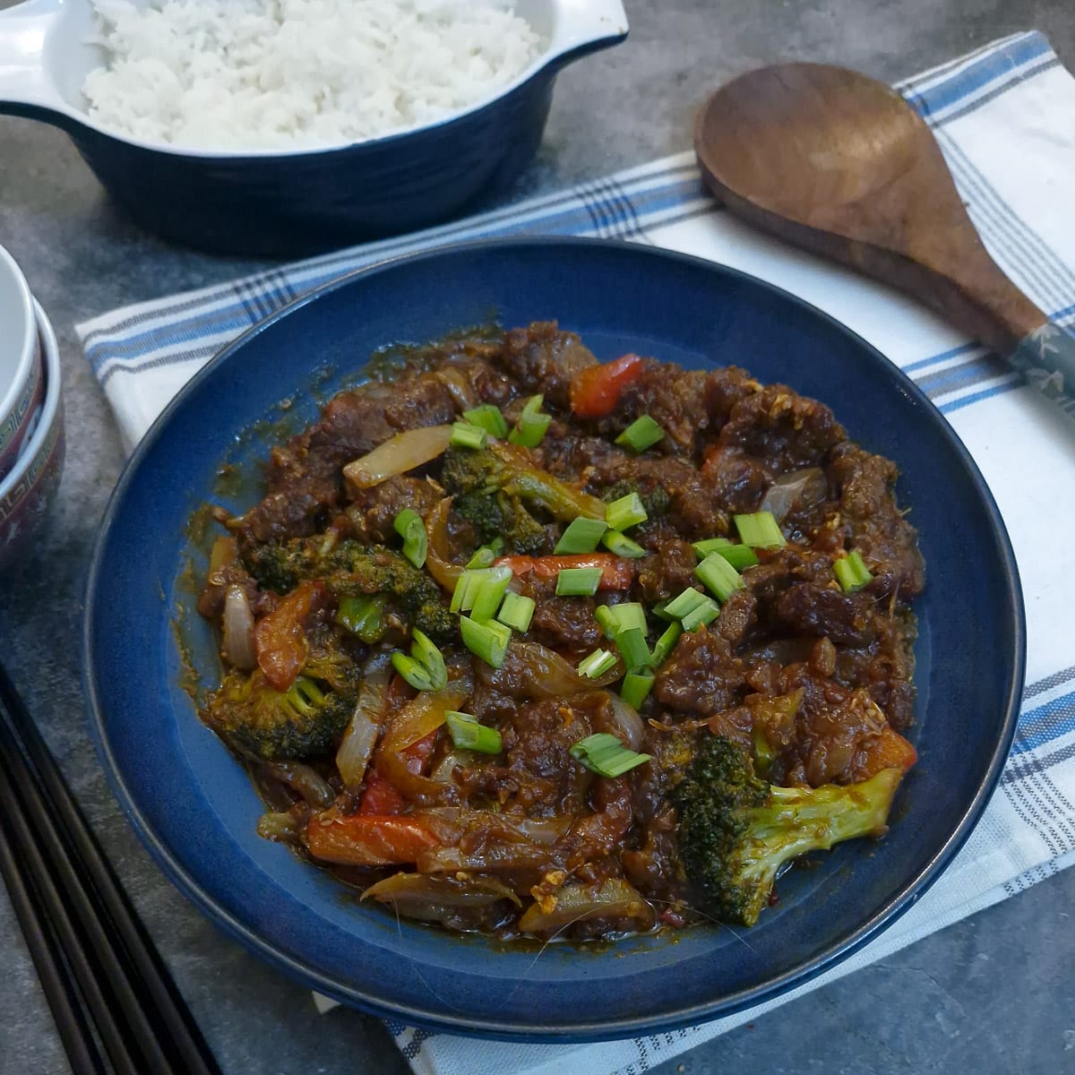 A blue serving dish of sweet chilli beef stir fry on a blue and white striped cloth. There is a large wooden serving spoon alongside.