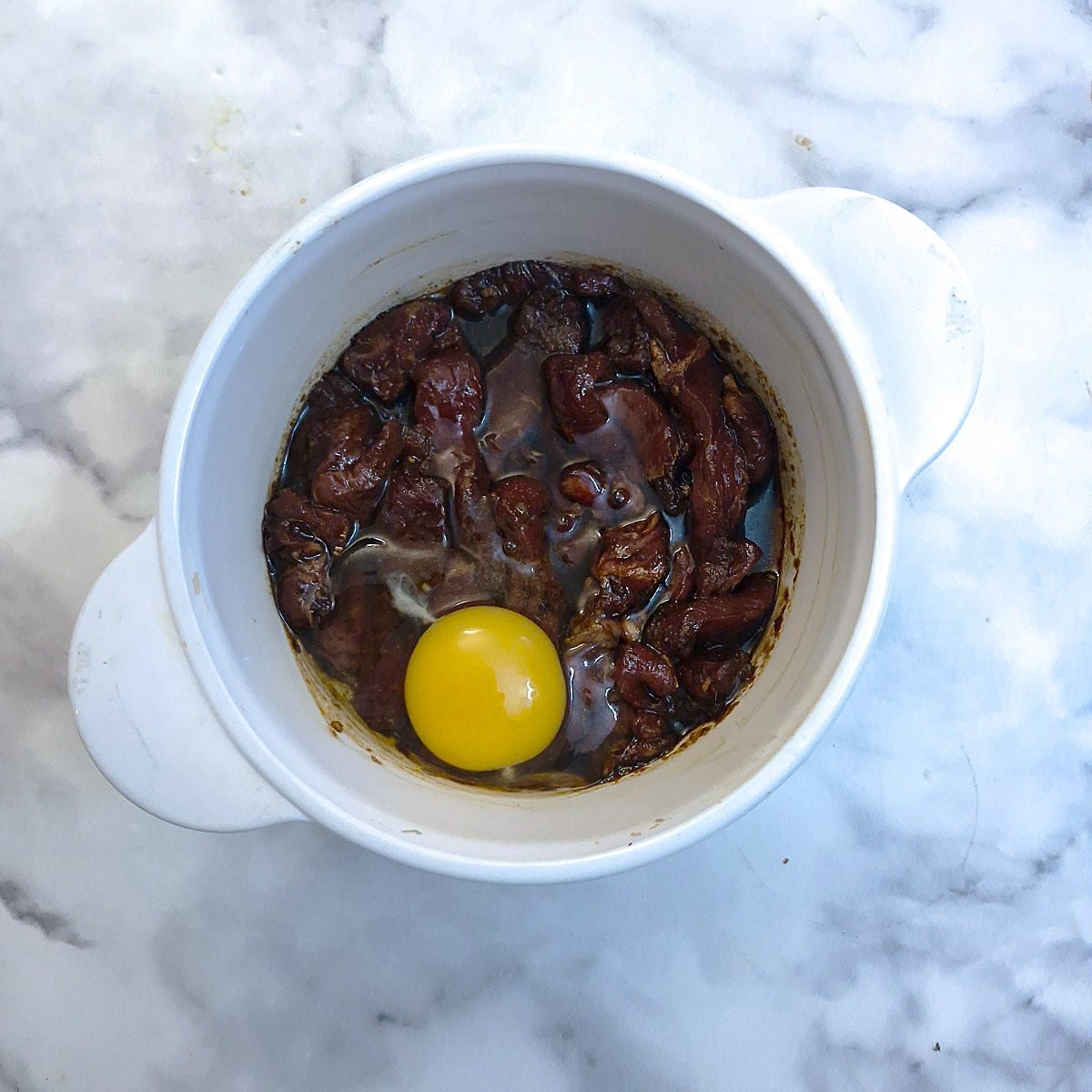 Beef strips in marinade in a white bowl. An egg has been broked into the bowl on top of the meat.
