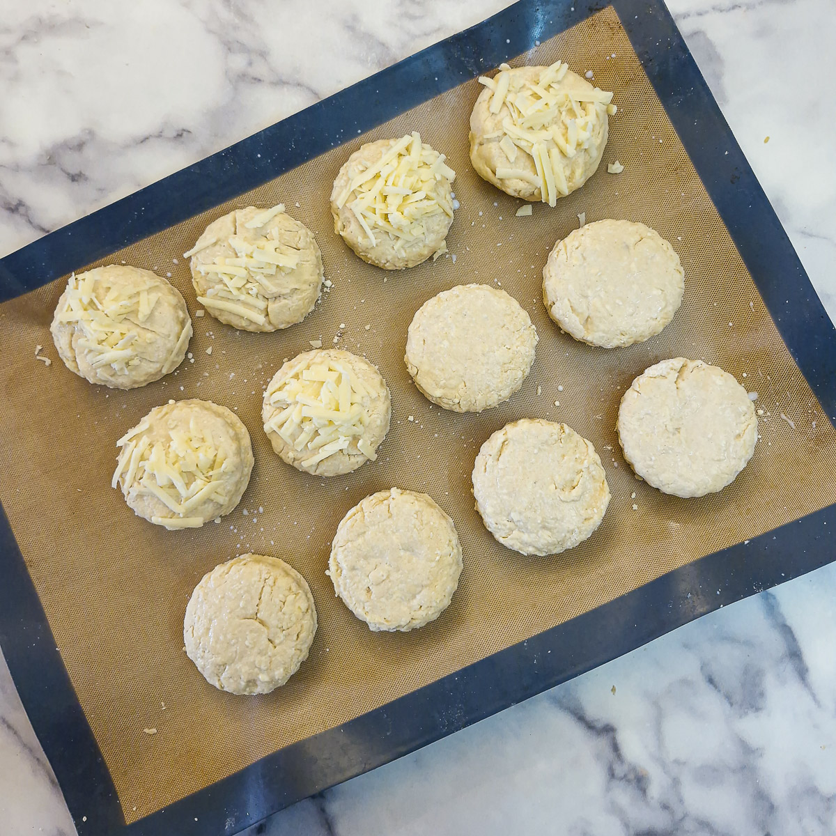 Unbaked cheese scones on a baking tray. Some of the scones have been topped with grated cheese.