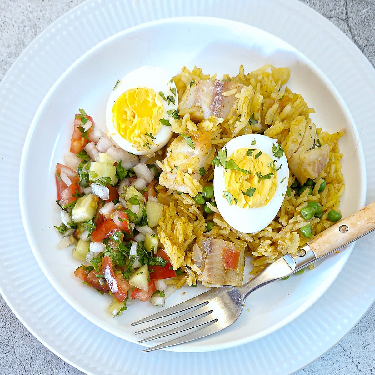 A plate of curried fish and rice pilaf with 2 halved hard-boiled eggs on top and a helping of kachumber salad on the side.