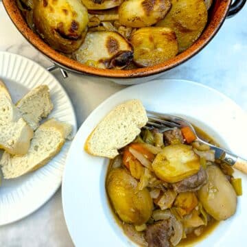 A deep white plate, filled with lamb potjie, with a slice of crusty bread on the side.