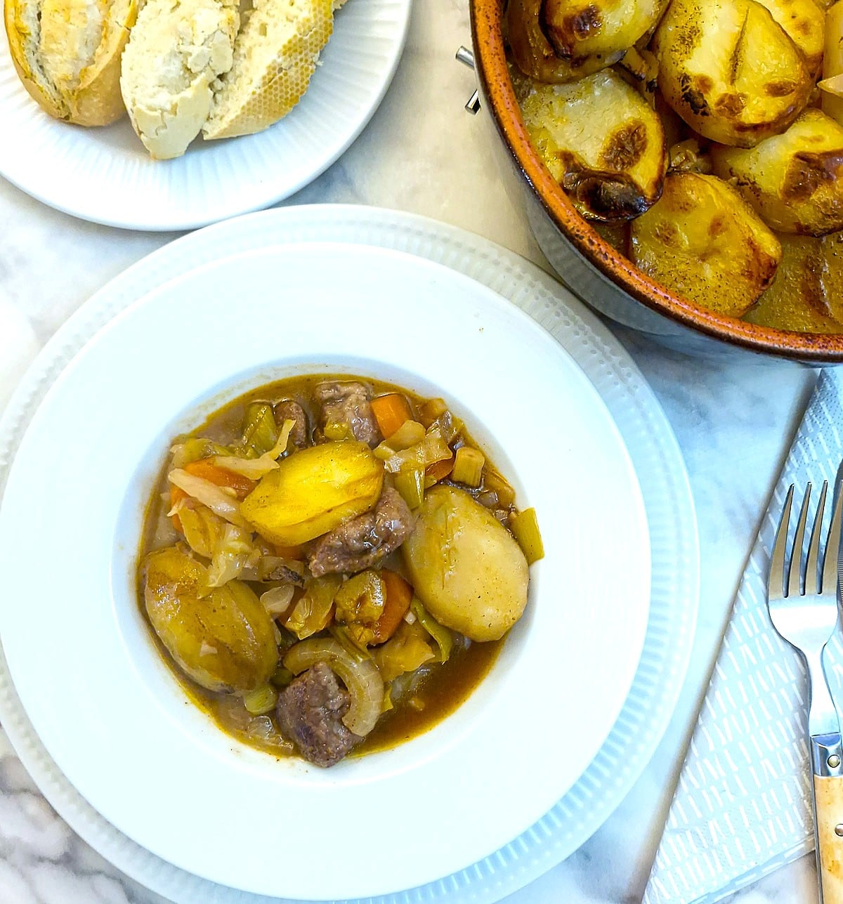 A white dinner plate with a helping of lamb potjie.  There is a plate of crusty bread and the serving dish of lamb stew alongside on the table.