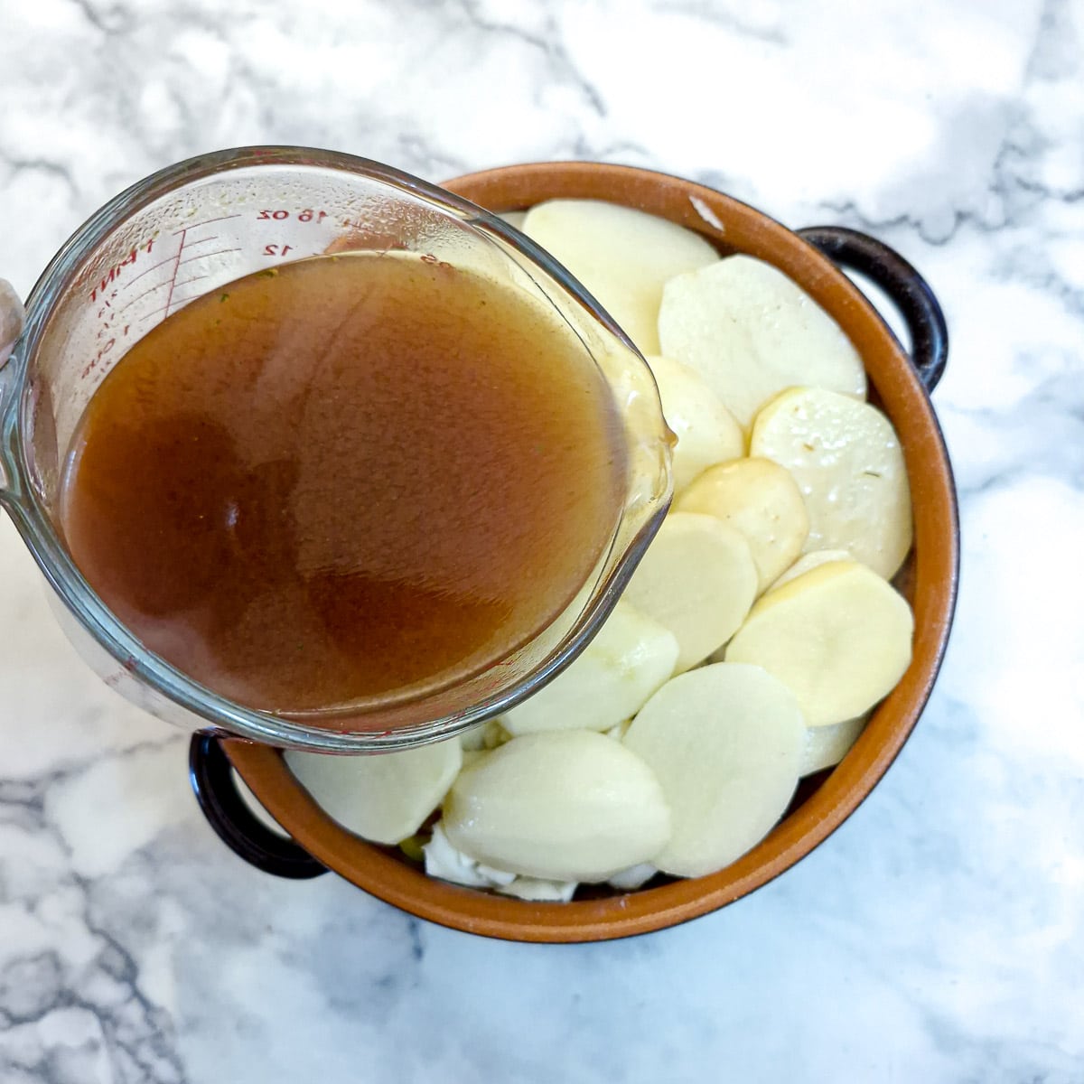 A glass jug of stock being poured into the casserole.