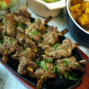 C;loseup of tandoori lamb skewers on a black cast iron plate resting on a wooden tray.