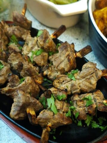 C;loseup of tandoori lamb skewers on a black cast iron plate resting on a wooden tray.