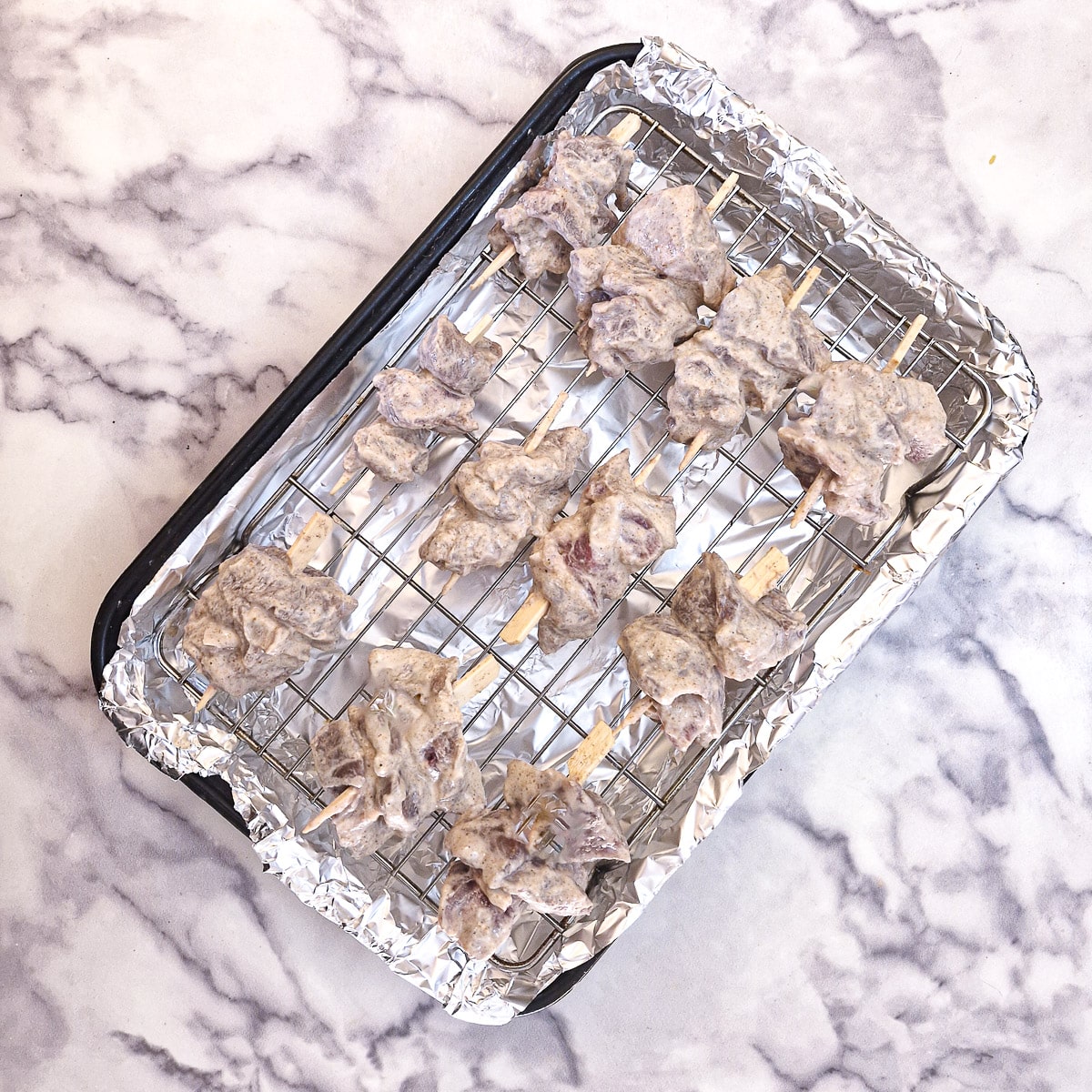 Uncooked tandoori lamb skewers on a rack set above a baking tray that has been lined with foil.