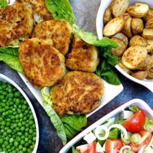A white oblong plate lined with lettuce with fish cakes arranged on top. There are dishes of peas, potatoes and lettuce and tomato salad to the side.