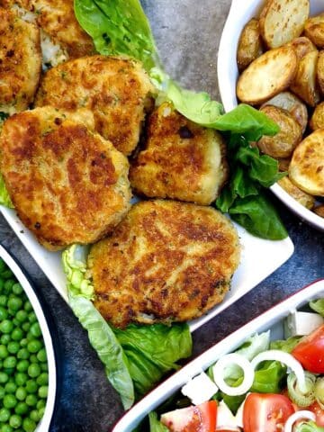 A white oblong plate lined with lettuce with fish cakes arranged on top. There are dishes of peas, potatoes and lettuce and tomato salad to the side.