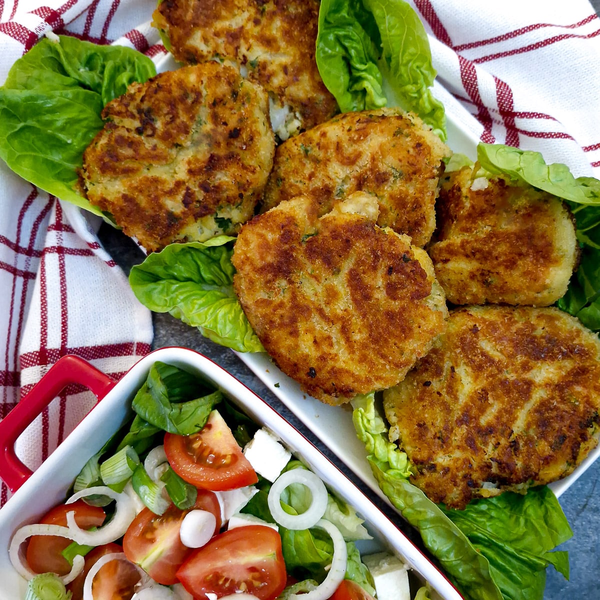 6 cooked fishcakes on a bed of lettuce on a serving plate next to a dish of tomato salad.