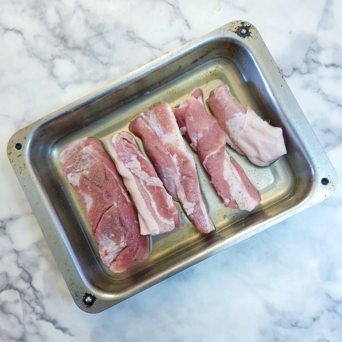 5 large pork belly slices in apple juice in a metal roasting tray.