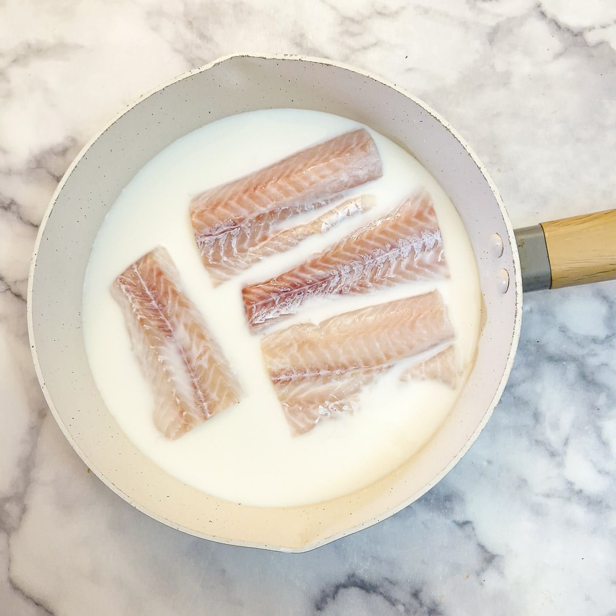 Pieces of white fish in milk in a frying pan ready to poach.