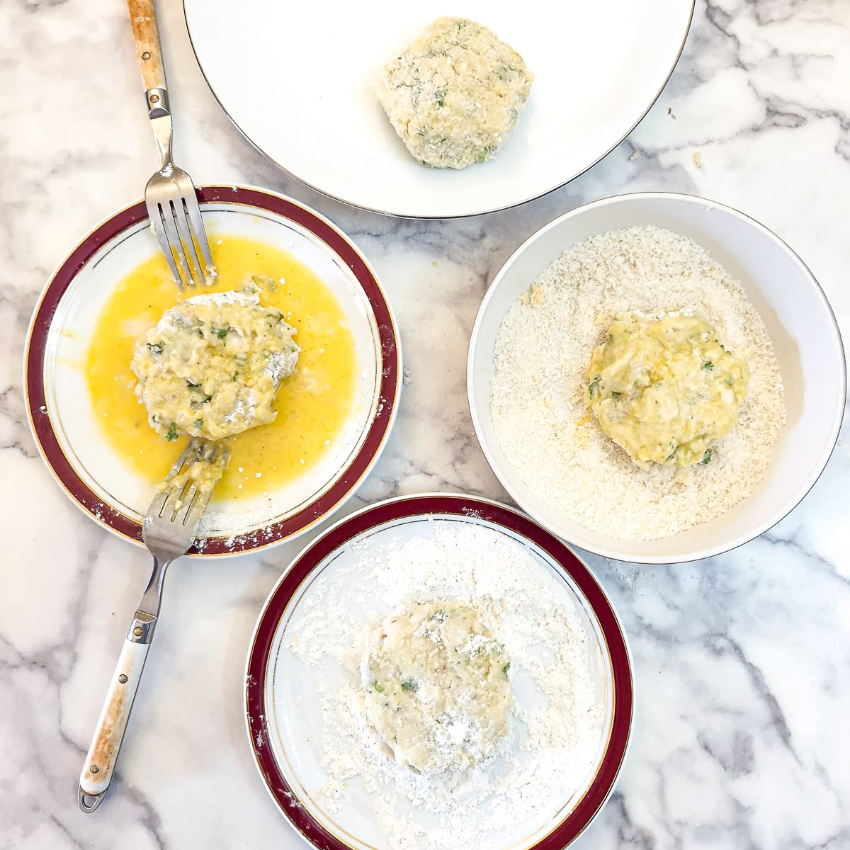 4 plates on a table. Once hollding flour, the second holding beaten egg, the third holding panko breadcrumb. There is a fish patty in each of them. The final plate holds a fully coated fish cake.