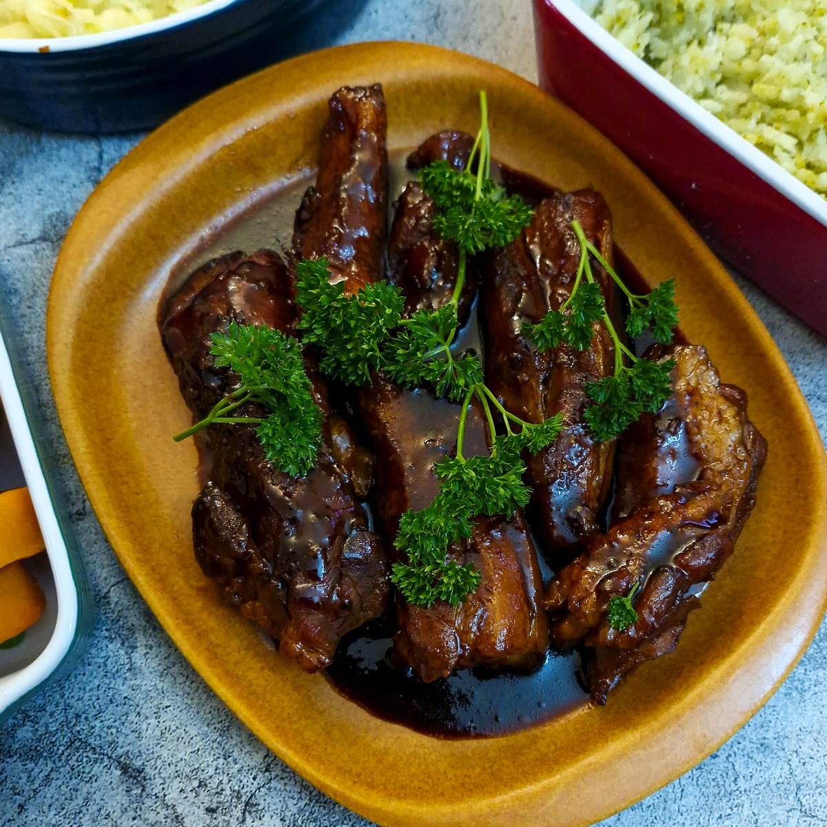 A plate of sticky pork belly rashers, garnished with parslesy and covered with maple glaze,
surrounded by vegetable side dishes.
