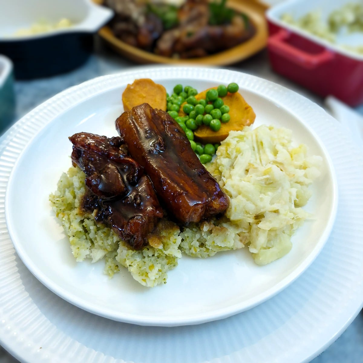2 glazed pork belly slices on a pile of vegetable mash on a white plate. There are also sweet potatoes and peas on the plate.