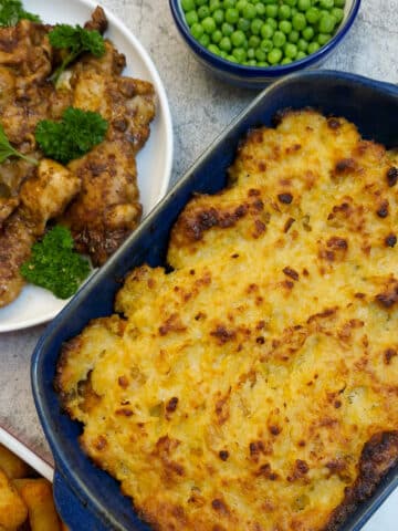 A blue serving dish of oven-baked butternut with corn topping. There is a plate of peri-peri chicken and a dish of peas alongside.