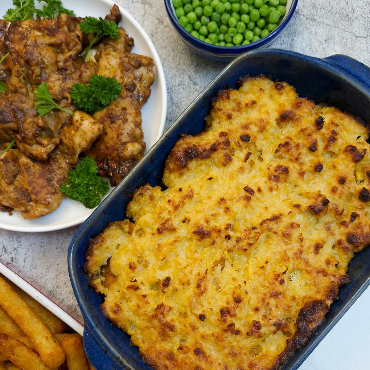 A blue serving dish of oven-baked butternut with corn topping. There is a plate of peri-peri chicken and a dish of peas alongside.