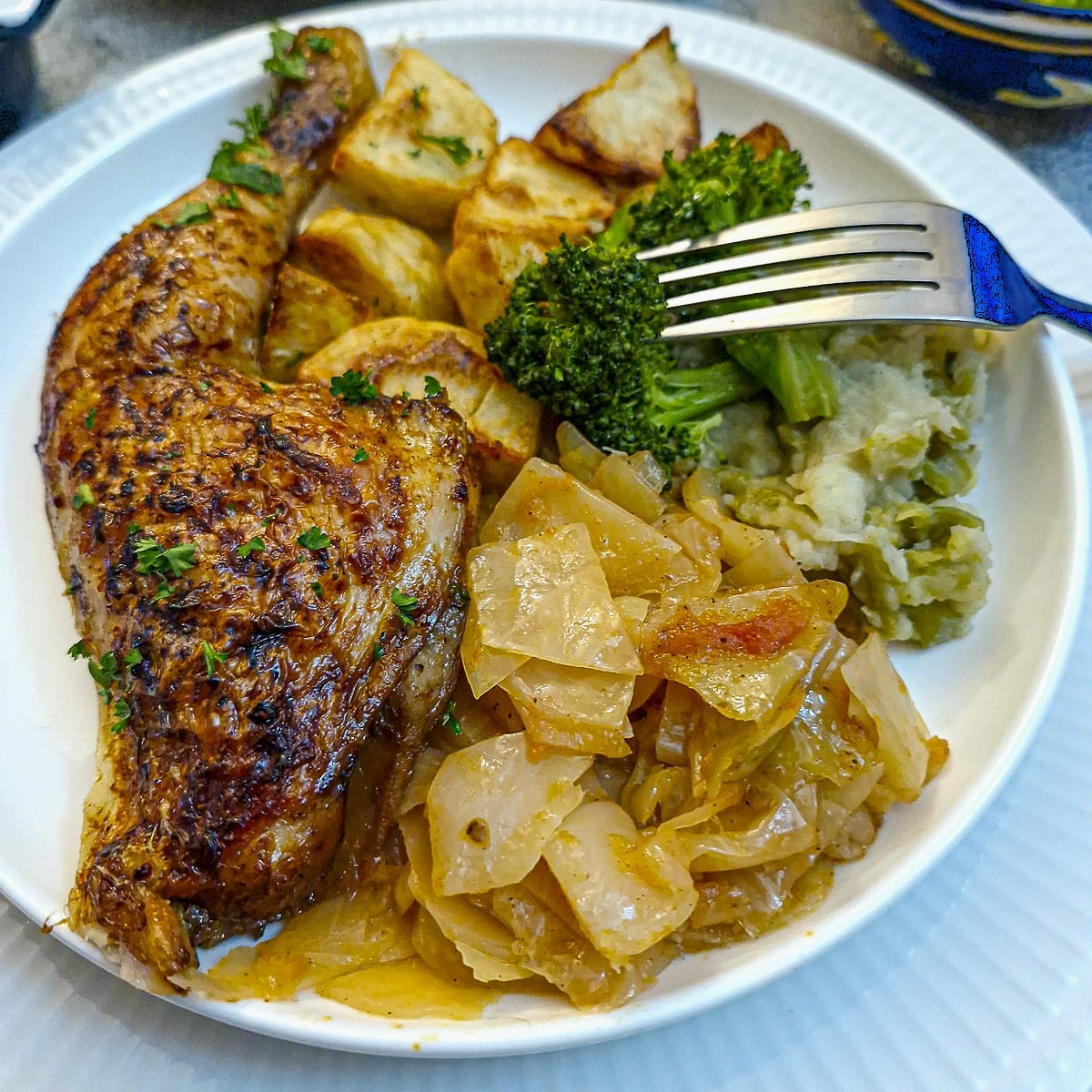 A browned, crispy chicken leg on a plate with tomato-braised cabbage, broccoli and roast potatoes.
