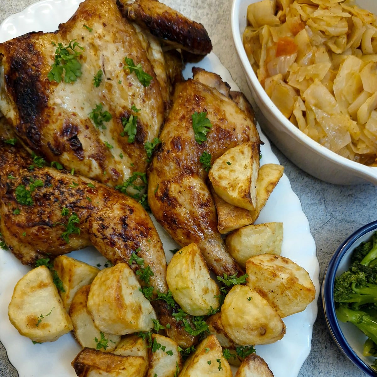 A honey and soy roasted chicken on a serving plate with roast potatoes. There are dishes of vegetables alongside.