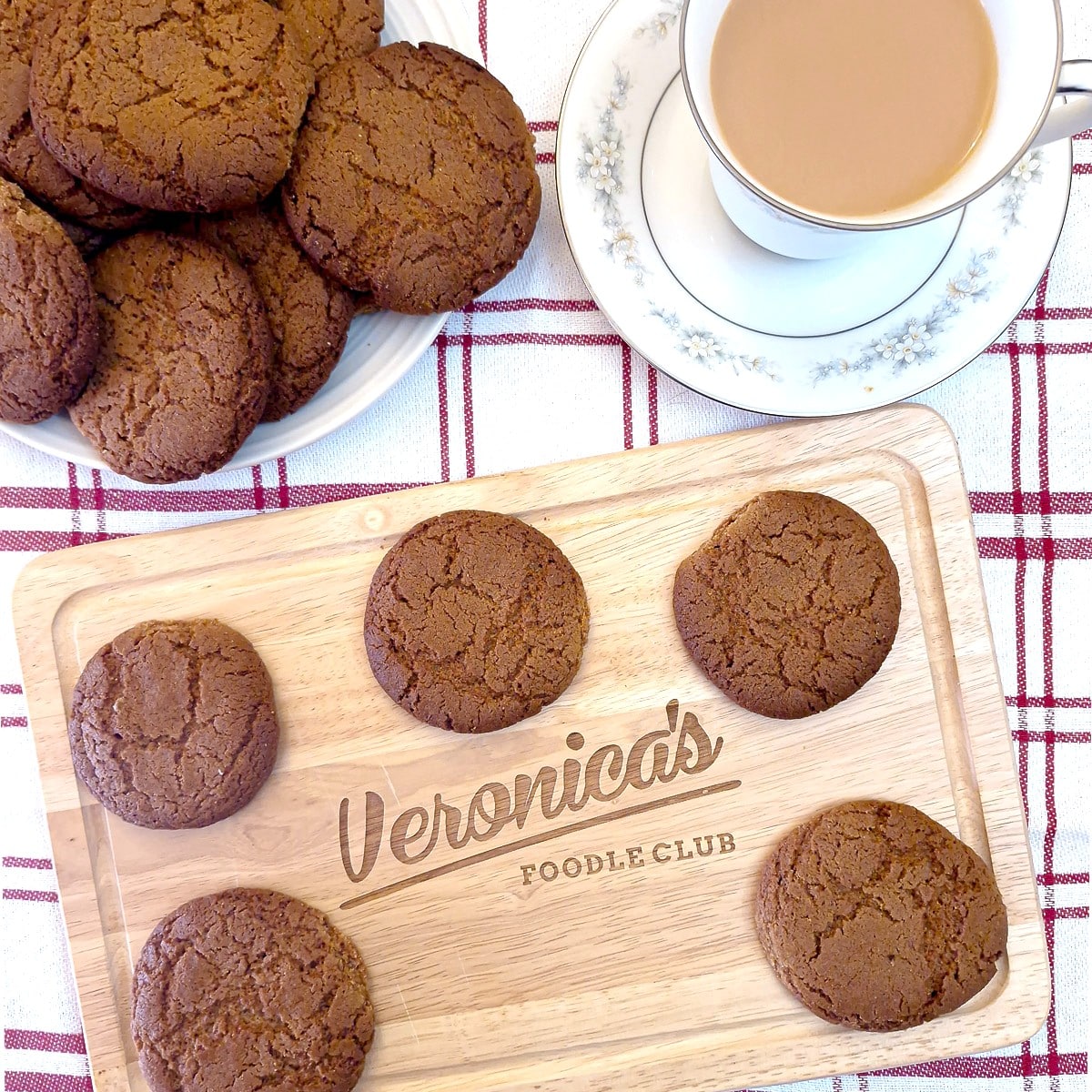 5 ginger snaps on a wooden board, with a plate of ginger snaps and a cup of tea in the background.