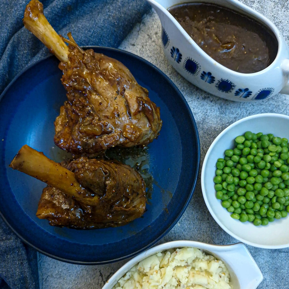 Two honey mustard lamb shanks on a plate with dishes of peas, cabbage and gravy alongside.