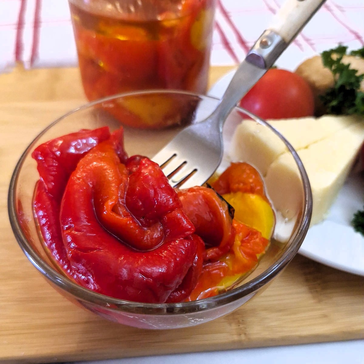 A glass dish holding roasted red peppers. There is a fork in the dish and a jar of peppers in the background.